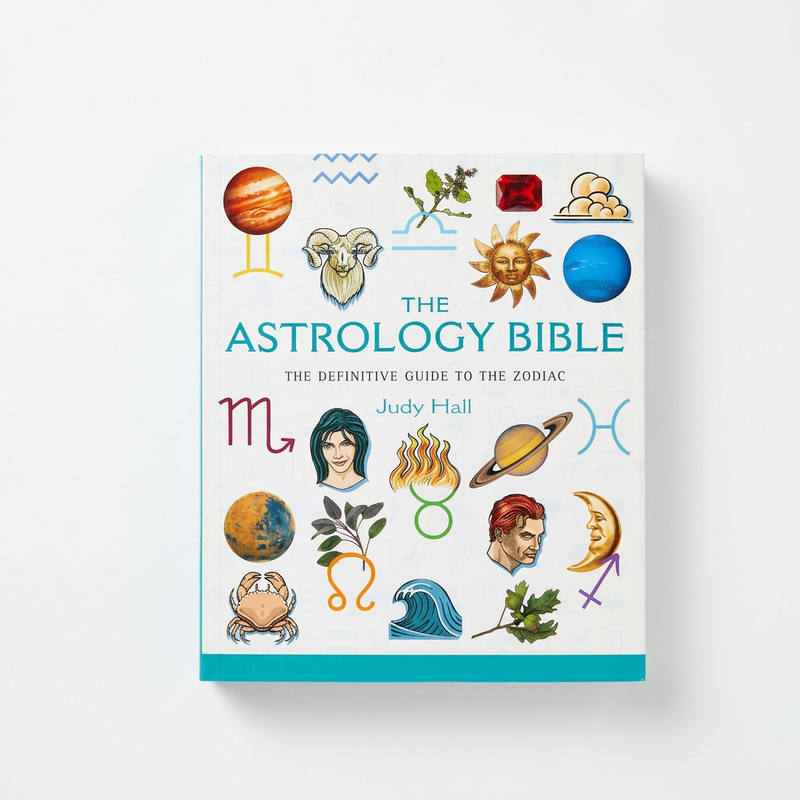 The Astrology Bible book by Judy Hall, featuring a white cover decorated with colorful astrological illustrations and zodiac symbols, shown against a light gray background.