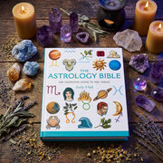 The Astrology Bible by Judy Hall is displayed on a rustic wooden surface surrounded by candles, crystals, herbs, and a black metal candle holder. The book cover features colorful astrological symbols and illustrations.