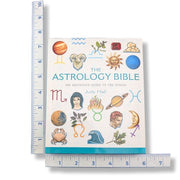 The Astrology Bible book by Judy Hall shown upright with ruler measurements along the left, bottom, and right edges to indicate size. The book cover is white with colorful zodiac and celestial illustrations, including a ram, sun, moon, planets, and astrological symbols.