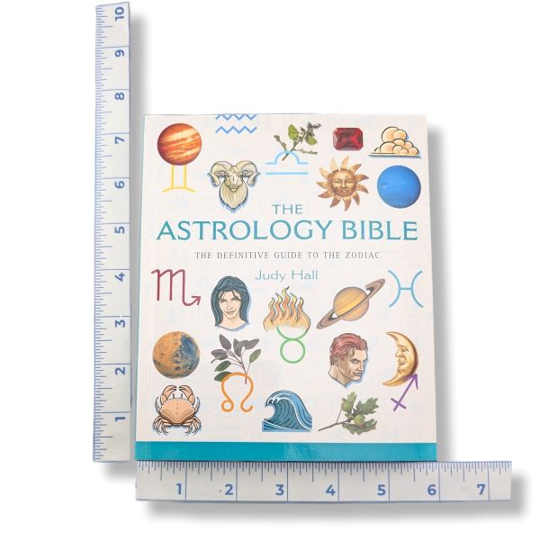 The Astrology Bible book by Judy Hall shown upright with ruler measurements along the left, bottom, and right edges to indicate size. The book cover is white with colorful zodiac and celestial illustrations, including a ram, sun, moon, planets, and astrological symbols.