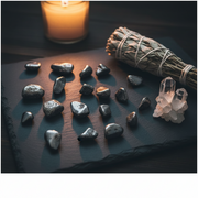 Mineral stones, crystal, and smudge stick on a dark surface with a candle.