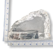 A raw, rough-cut clear selenite crystal specimen with visible internal inclusions and natural fracture lines, shown with a blue ruler on each side to indicate its dimensions against a white background.