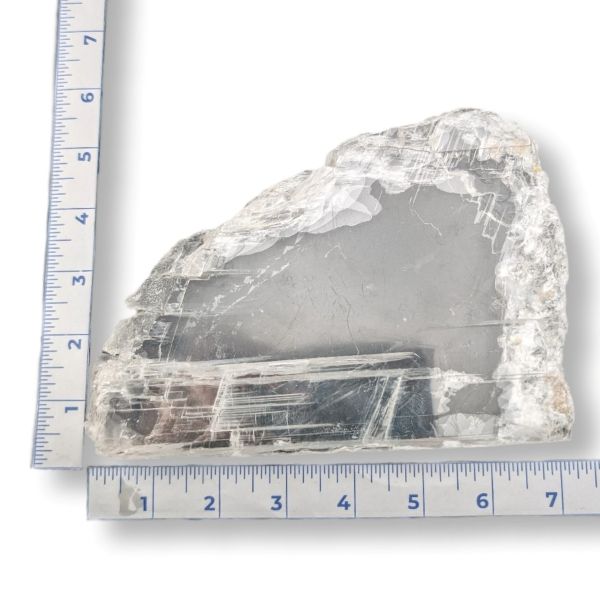 A raw, rough-cut clear selenite crystal specimen with visible internal inclusions and natural fracture lines, shown with a blue ruler on each side to indicate its dimensions against a white background.
