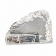 A raw, rough-cut clear selenite specimen showing layered tabular crystal structures and internal gray smoky areas, photographed on a clean white background.