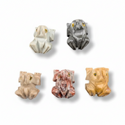 Five small frog-shaped jewelry pieces or charms in different stone-like colors, arranged on a plain white background.