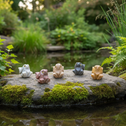 Five small frog figurines in different earthy colors are arranged on a mossy rock beside a pond, with a blurred garden background.