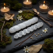Two carved white selenite wand pieces displayed on a dark slate surface with moss, dried leaves, small white flowers, and lit candles in the background.