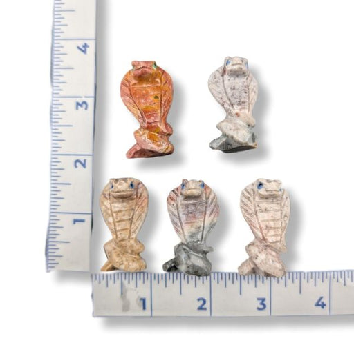 Five small carved cobra figurines in various earthy tones, arranged on a white background with a ruler on each side for scale.