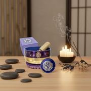 A decorative purple and gold singing bowl set with a matching lid, wooden striker, and small box, arranged on a wooden table beside spa stones, lit candles, incense, and dried flowers in a calm indoor setting.