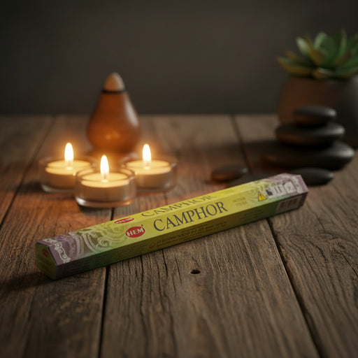 Pack of HEM Camphor incense sticks on a wooden surface with 3 candles in the background with rocks  and plant