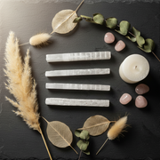 Four white selenite crystal sticks arranged vertically on a dark slate surface, styled with dried pampas grass, eucalyptus leaves, pink stones, and a white pillar candle.