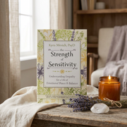 A book titled 'the Strength of Sensitivity' by Kyra Mesich, PsyD stands on a rustic wooden table in a cozy room setting. The book cover features a cream background with green and purple botanical illustrations.