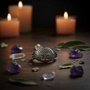 Silver ring with tree of life design on a table with 3 candles in the background and the ring surrounded by quartz, amethyst and dried herbs.