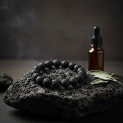A black lava stone bead bracelet arranged in a circle on a dark volcanic rock, with an amber dropper bottle and green leaves in the background against a dark smoky backdrop.