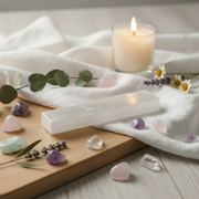 A rectangular white selenite crystal ruler 6 inches length resting on a wooden board, surrounded by pastel tumbled stones, a lit candle in a glass jar, white flowers, and green eucalyptus leaves on a white fabric surface.