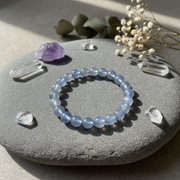 A light blue beaded gemstone bracelet arranged in a circle on a smooth gray stone, surrounded by clear crystal points, a purple crystal, and dried white flowers in soft natural sunlight.
