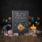 Book titled 'The Little Book of Crystals' surrounded by crystals and a candle on a wooden surface with blurred lights in the background.