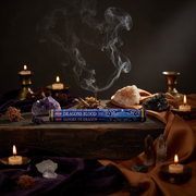 A box of Wajra Magic Incense labeled 'Dragons Blood' sits on a dark wooden table surrounded by lit candles, crystals, and draped purple and brown fabric, with smoke rising above.