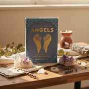A book titled 'A Little Bit of Angels' by Elaine Clayton stands upright on a wooden table surrounded by crystals, dried lavender, a sage bundle, and a small terracotta candle holder in a warm, sunlit setting.