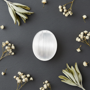 White Selenite palm stone on a grey background surrounded by herbs