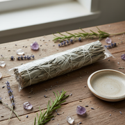 A bundled bundle of dried herbs tied with string, wrapped in clear plastic, resting on a rustic wooden table with rosemary sprigs, lavender flowers, amethyst crystals, and a small ceramic dish.