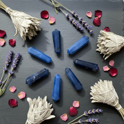 Six blue crystal points arranged in a circle on a dark slate background, surrounded by dried white sage bundles, lavender sprigs, and scattered pink and red flower petals.