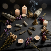 A styled arrangement of spiritual and ritual items on a dark slate surface, including white and cream candles, two white sage smudge bundles, a brass incense holder with a smoking stick, several gray crystal points, dried lavender and white flowers, and dried roses against a dark background with soft bokeh lights.