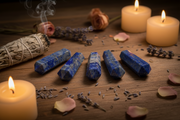 Five blue lapis lazuli crystal points arranged in a row on a wooden surface, surrounded by lit candles, rose petals, dried lavender, a sage bundle, and a rose.