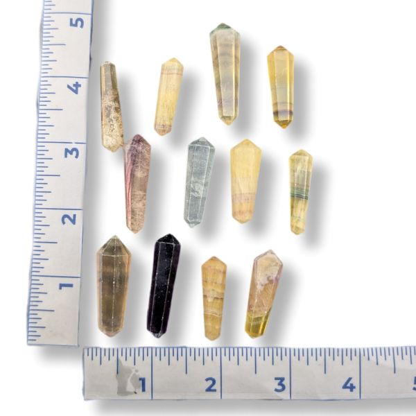 Nine small pointed fluorite points in various natural colors and patterns, arranged on a white background with ruler markings showing their size.