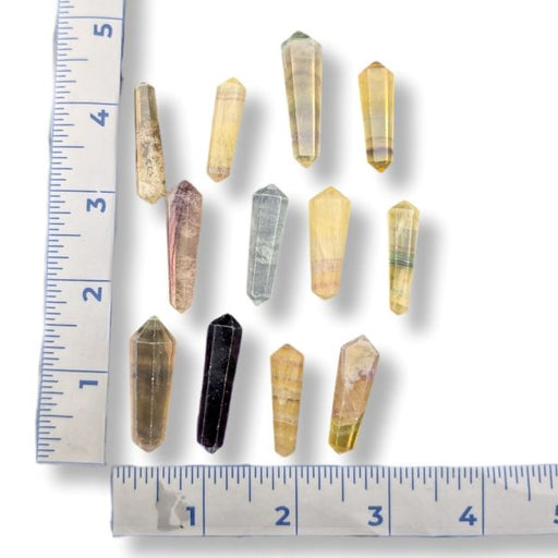 Nine small pointed fluorite points in various natural colors and patterns, arranged on a white background with ruler markings showing their size.