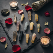 A flat-lay arrangement of various fluorite points in yellow, purple, white, and black, styled with red rose petals, dried leaves, and two lit tealight candles on a dark slate background.