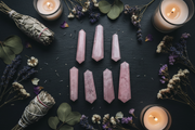 Six pink crystal points arranged in two rows on a dark slate surface, surrounded by dried lavender, white flowers, green leaves, and two lit candles.