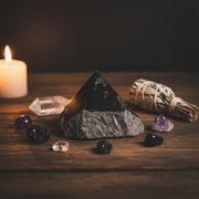 Pyramid-shaped stone, crystals, and a lit candle on a wooden surface with a dark background.
