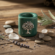 A tealight candle in a glossy dark green cylindrical holder with a silver tree of life emblem on the front, lit and placed on a rustic wooden surface surrounded by stones, lavender, and dried leaves.