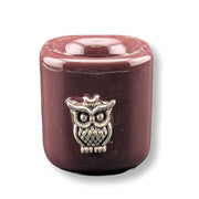 A small cylindrical candle in a glossy dark reddish-brown finish, featuring a silver-toned metal owl charm on the front, set against a plain white background.