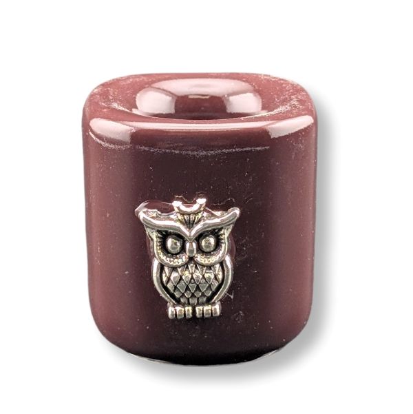 A small cylindrical candle in a glossy dark reddish-brown finish, featuring a silver-toned metal owl charm on the front, set against a plain white background.