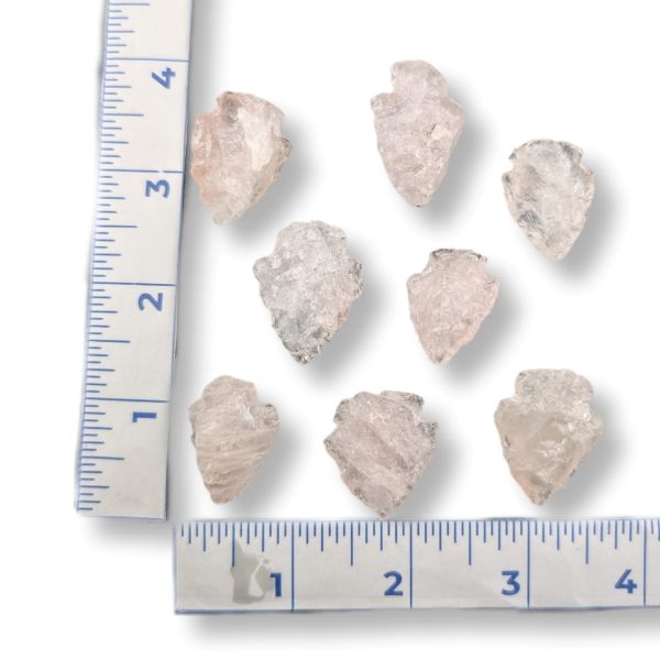 Seven raw, rough translucent stone rose quartz arrowheads arranged on a white background with a blue ruler on the left and bottom for size reference.