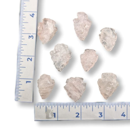 Seven raw, rough translucent stone rose quartz arrowheads arranged on a white background with a blue ruler on the left and bottom for size reference.