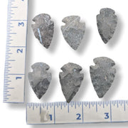Six dark gray stone arrowhead tools arranged on a white background with ruler measurements along the left and bottom edges for scale.