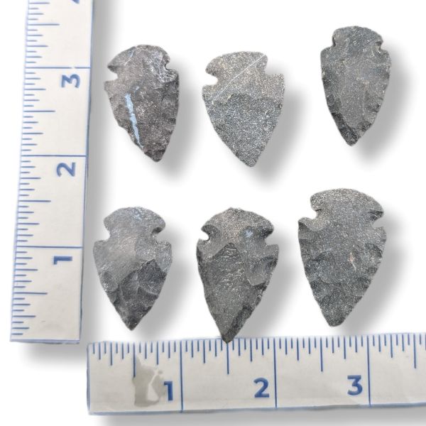 Six dark gray stone arrowhead tools arranged on a white background with ruler measurements along the left and bottom edges for scale.