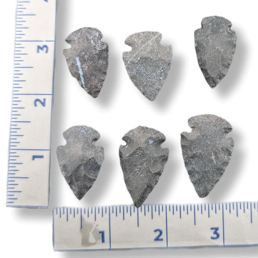 Six dark gray stone arrowhead tools arranged on a white background with ruler measurements along the left and bottom edges for scale.