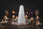 A white selenite tower crystal stands on a dark wooden surface, surrounded by pink rose quartz stones, a sage smudge bundle, and small burning tealight candles against a dark blurred background.