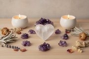 A heart-shaped selenite centerpiece surrounded by purple amethyst clusters, dried flowers, and two lit white candles on a light wood surface.
