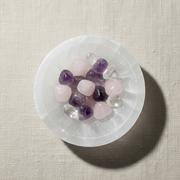 A top-down view of polished tumbled crystals, including pink rose quartz, purple amethyst, and clear quartz, arranged in a circular white selenite 6cm bowl on a textured beige linen surface.