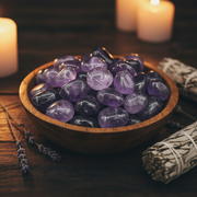 Purple fluorite tumbled stone in a bowl on a table with candles and herbs around the bowl