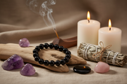 Black beaded bracelet on a wooden stick with purple crystals, candles, and sage on a textured surface.