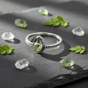 Silver ring with a green gemstone on a grey slate piece with leaves and crystals scattered around.