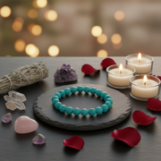 A turquoise beaded bracelet displayed on a dark slate coaster, surrounded by candles, rose petals, crystals, and a smudge bundle on a dark tabletop with a warm bokeh background.