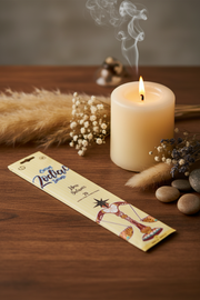 A packaged incense stick set labeled 'Zodiac incense' lies on a wooden tabletop beside a lit cream pillar candle, dried grasses, small white flowers, and smooth stones. Smoke curls around the candle flame.