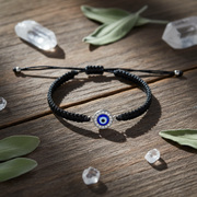 Black braided bracelet with a blue evil eye charm on a wooden surface with crystals and leaves.